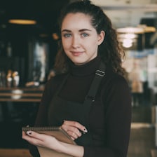 Une restauratrice en tablier, carnet en main, dans sa salle.